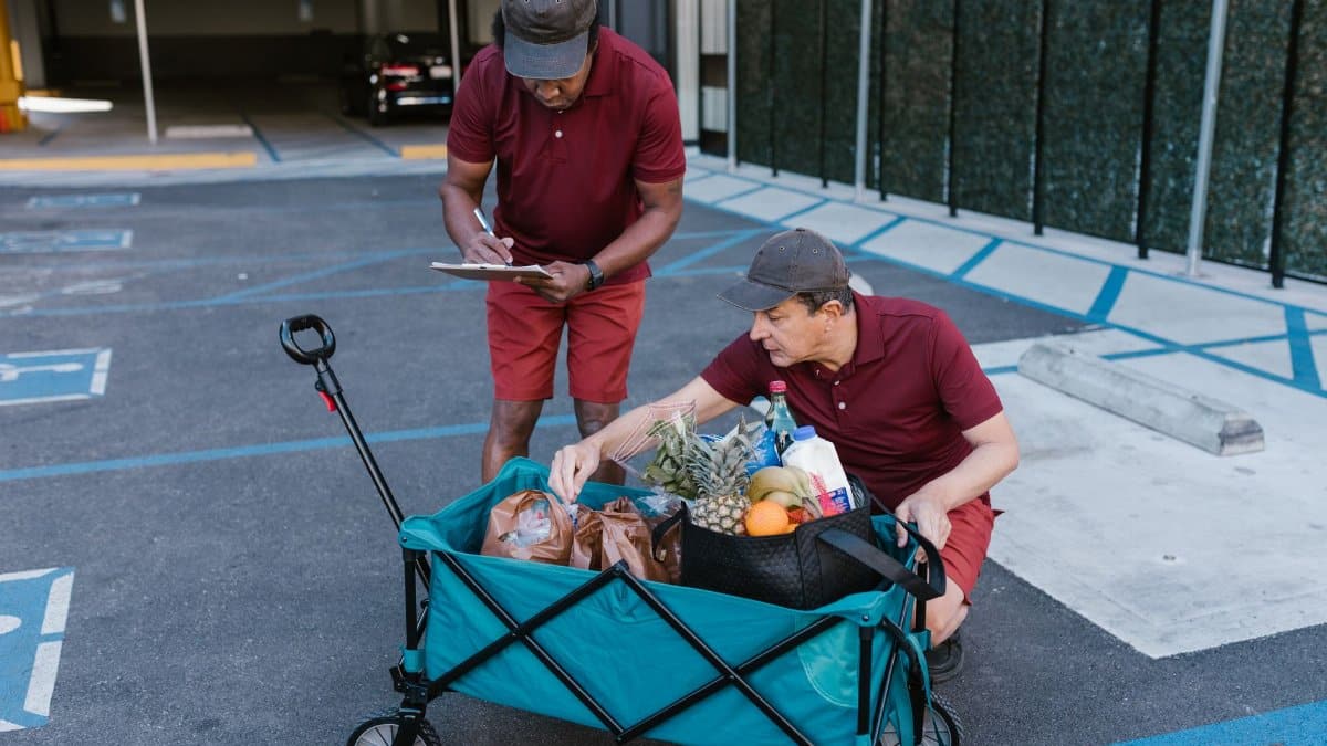 Two men in red uniforms delivering groceries with a push cart in a parking area.