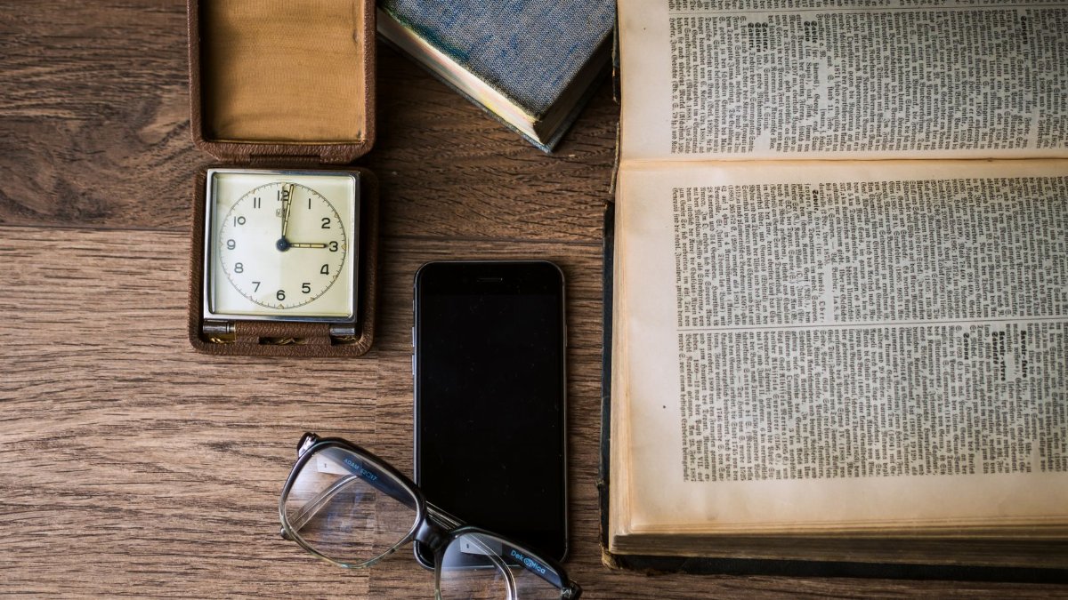 Flat lay of a vintage clock, open book, smartphone, and eyeglasses on a wooden table.