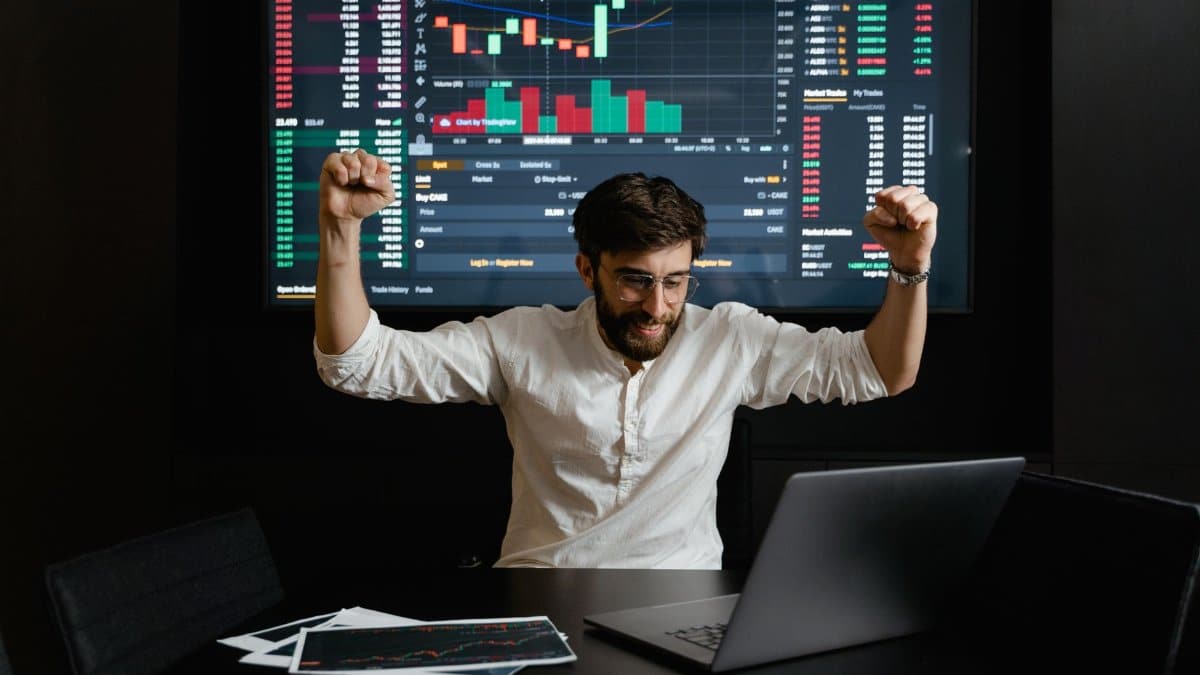 Businessman celebrates stock market success with hands raised in excitement at a trading desk.