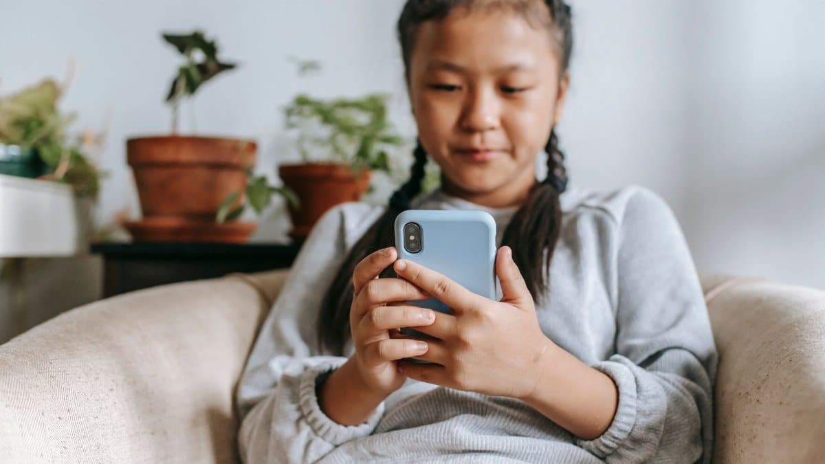 Focused Asian girl in casual clothes sitting on cozy armchair and browsing social media on smartphone