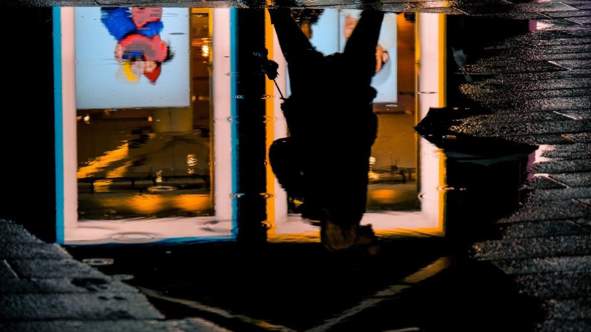 A silhouette of a person is reflected in a water puddle, capturing urban lights and textures.