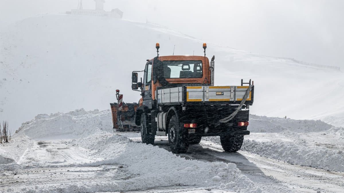 A snow plow truck clearing a snowy road near a hilltop in Andorra under cloudy skies.