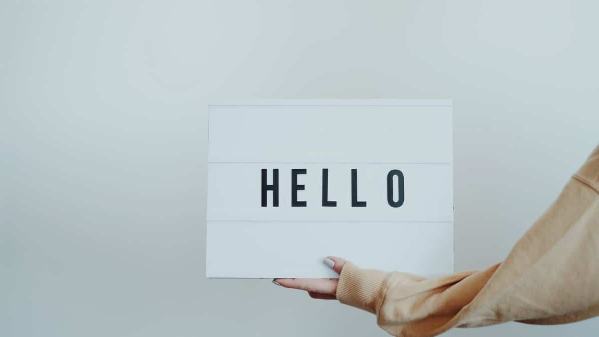 A hand holds a white sign displaying the word 'Hello' against a plain background.