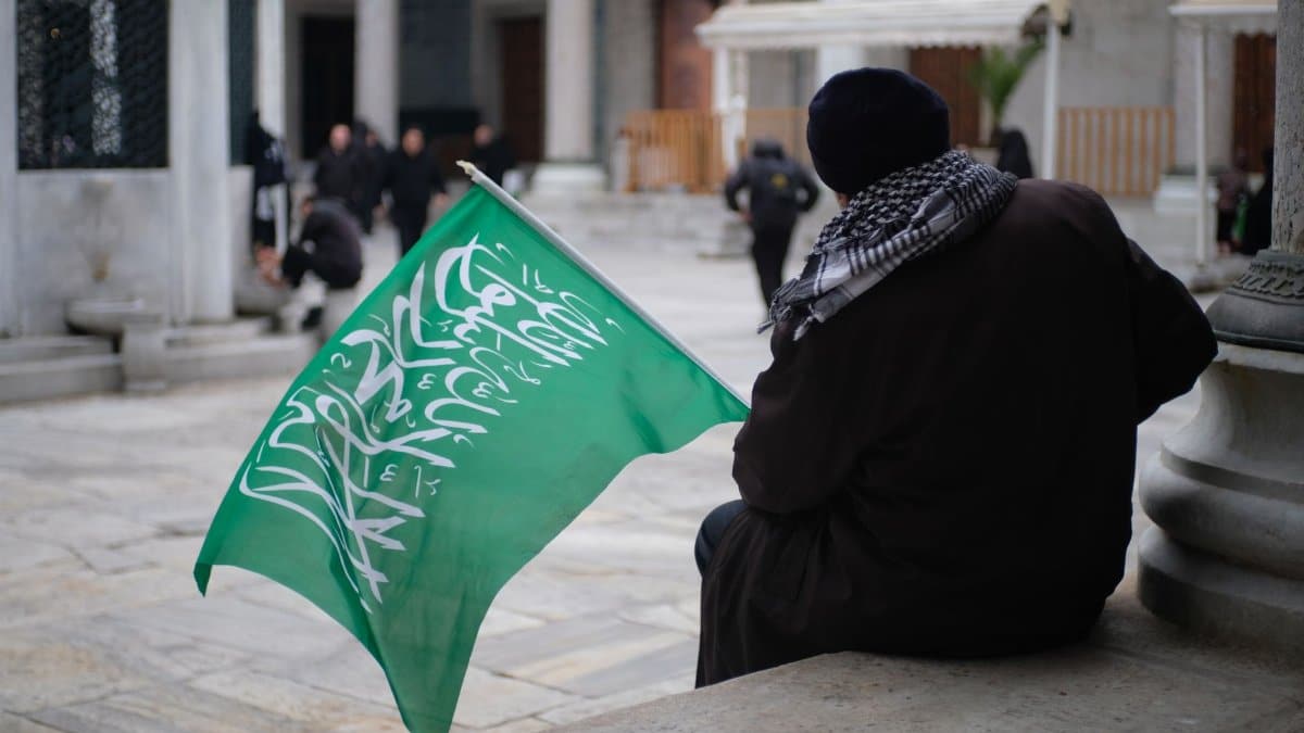 A man sits holding a green flag with Arabic script on a city street.