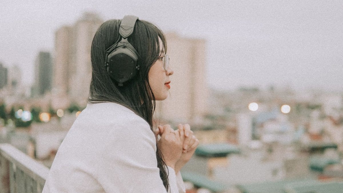 Side view of a woman in headphones and eyeglasses, enjoying a cityscape from a rooftop.