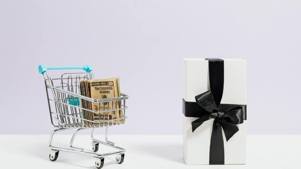 Shopping cart filled with dollar bills next to a black ribbon gift box, symbolizing commerce and presents.