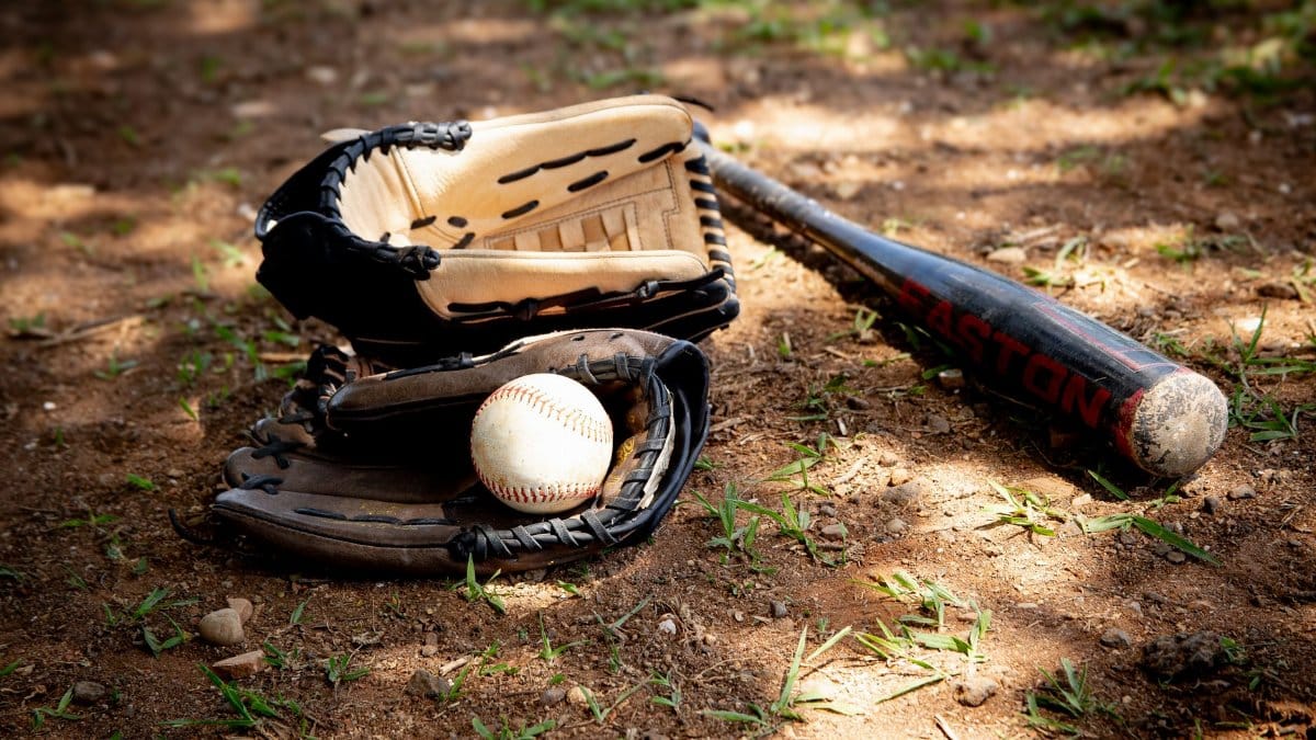 Close-up of baseball equipment including ball, bat, and gloves on a rural dirt ground.