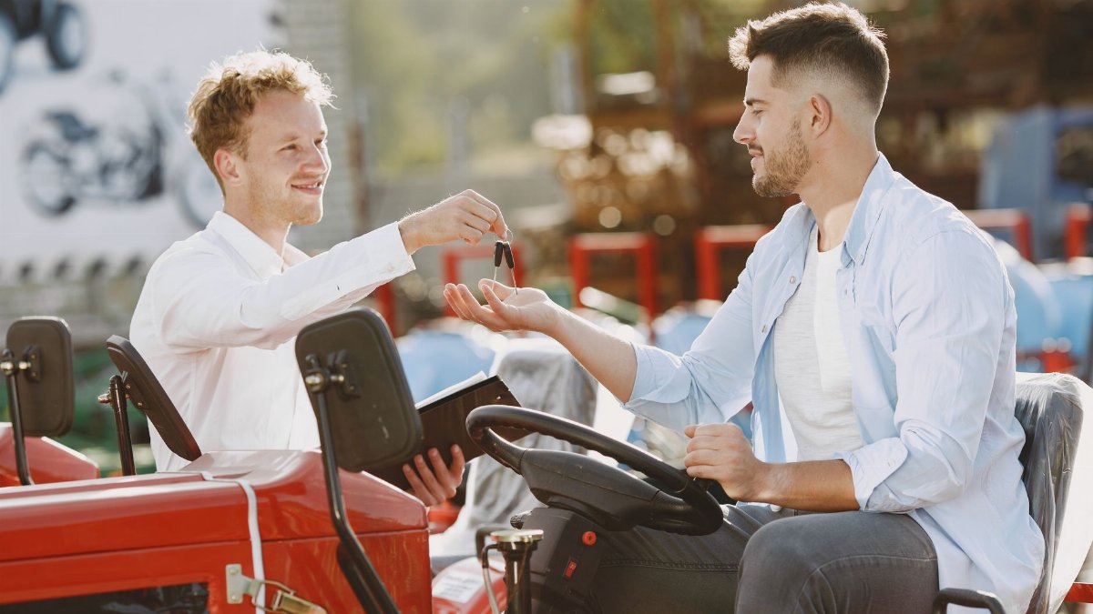 Men exchanging keys with a handshake by a red tractor in a bright, outdoor setting.