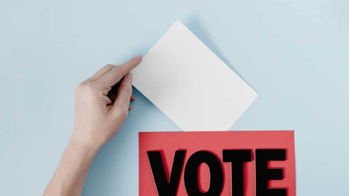 A hand places a ballot into a red box with the word VOTE against a blue background.