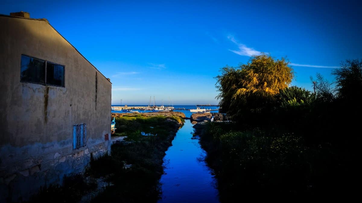 Serene coastal scene with a canal flowing into the sea, bordered by lush trees and an old building under a bright blue sky.