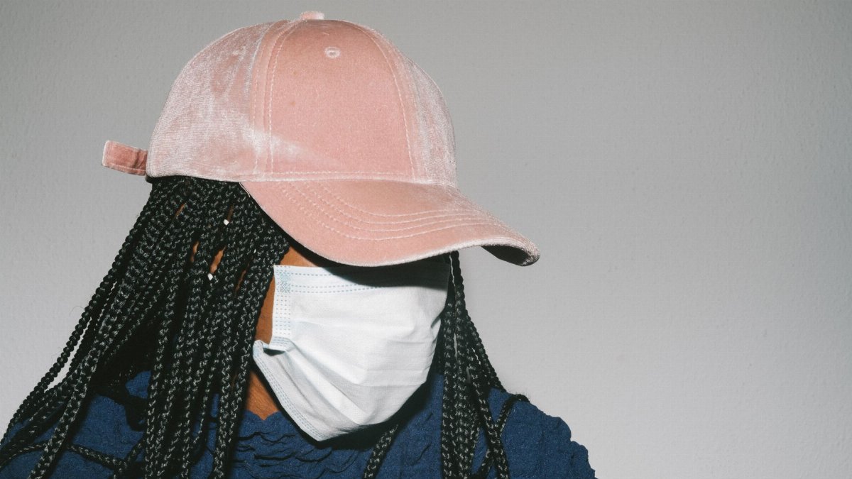 Faceless African American female in casual outfit and pink cap and protective mask on gray background