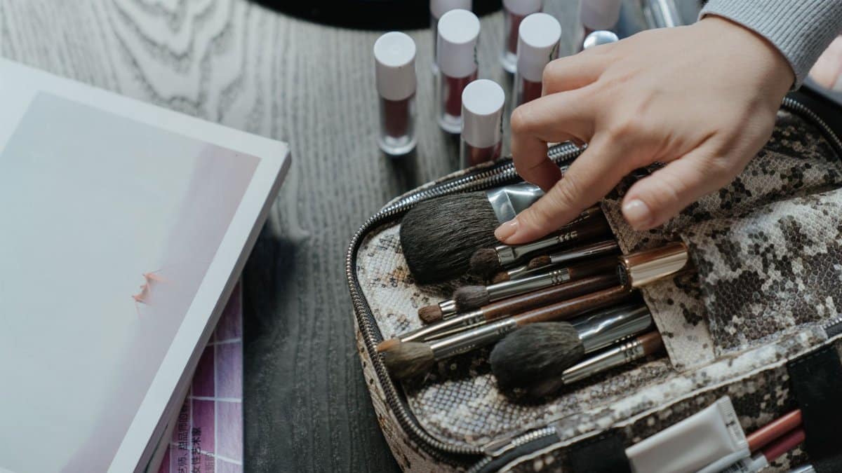 A hand arranging makeup brushes in a case surrounded by cosmetics in a flat lay setup.