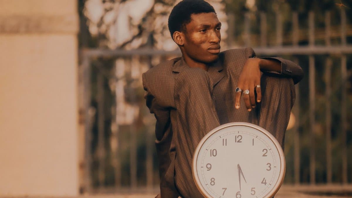 A contemplative man in a suit sits outdoors holding a large clock, embodying the passage of time.