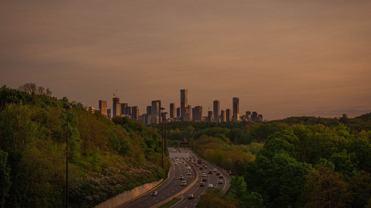 Aerial shot of city skyline with highway and forest during sunset.