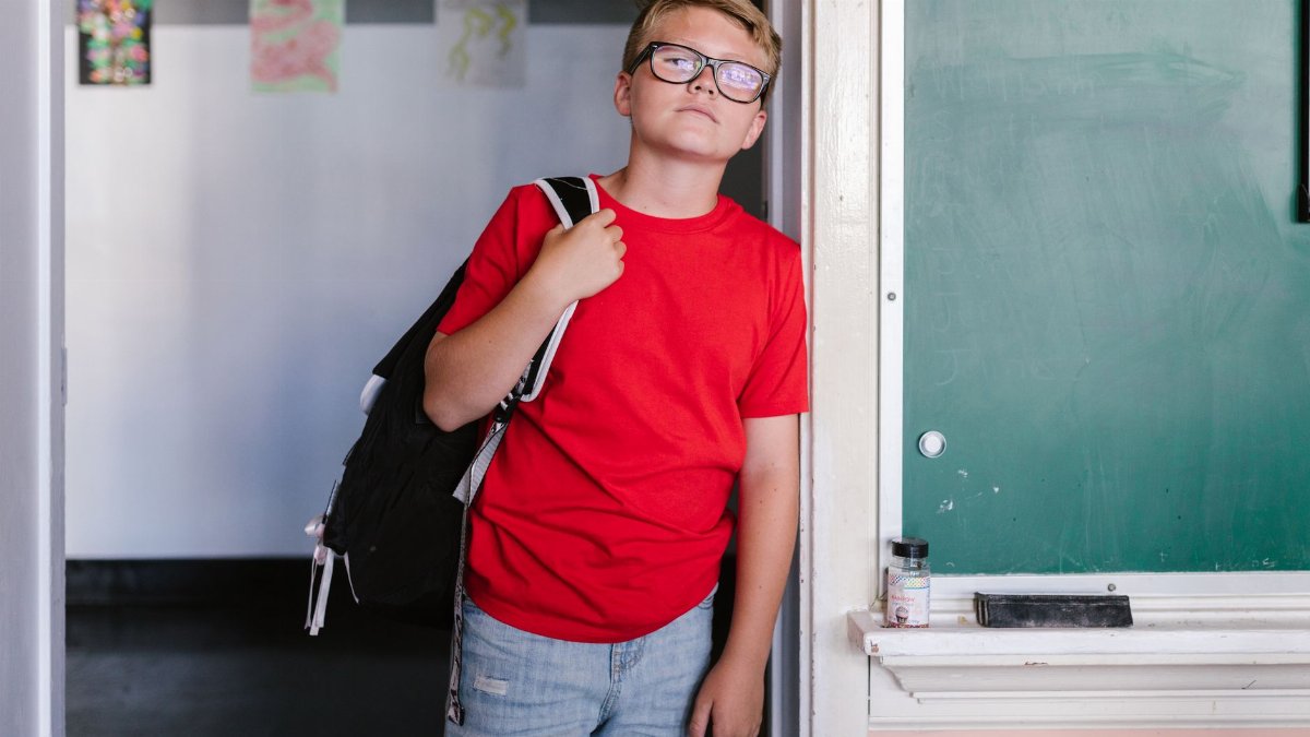 Boy in red shirt and jeans standing by a chalkboard in a classroom.