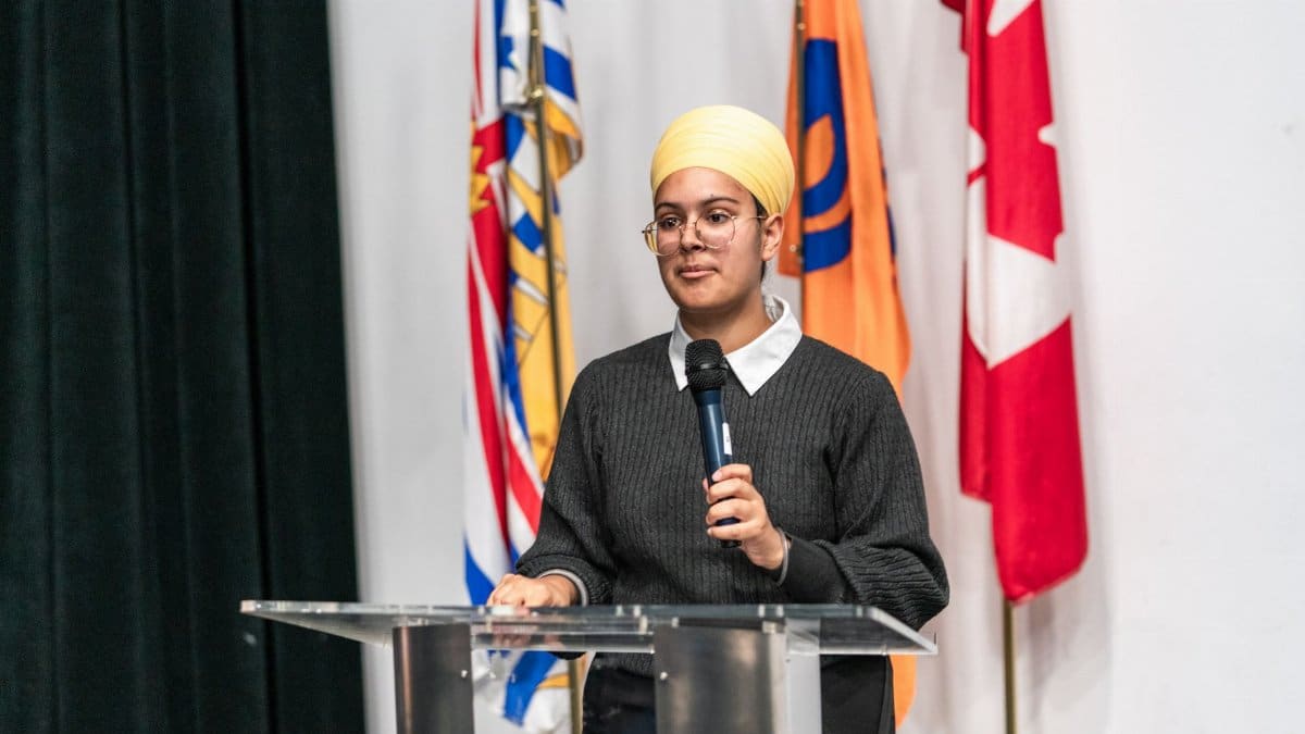 A young woman wearing a turban speaks at a podium with international flags in the background.