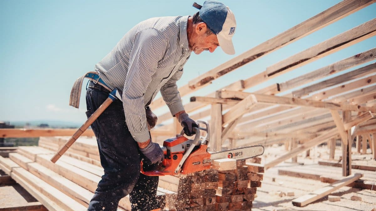 Skilled carpenter using a chainsaw to cut timber at an outdoor construction site under clear skies.