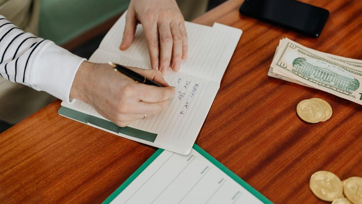 Person writing in a notebook on a wooden table with cash, coins, and a phone nearby.