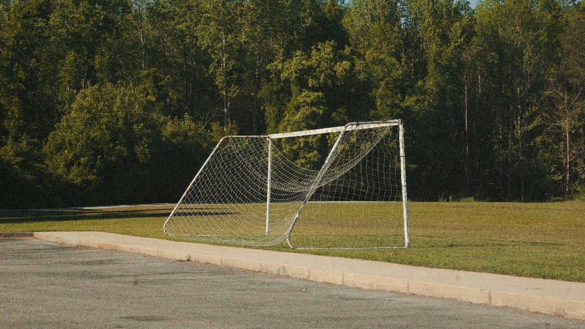 A rural soccer field featuring a bent goalpost with lush green trees in the background.