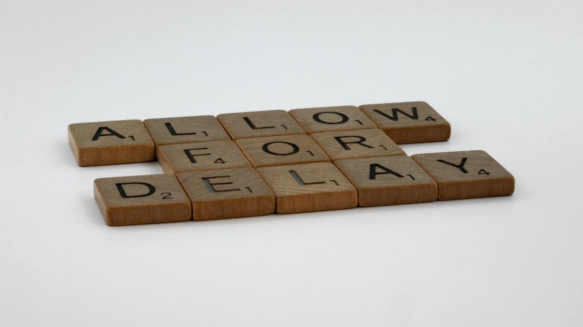 Wooden Scrabble tiles arranged on a white surface spelling 'Allow for Delay'.
