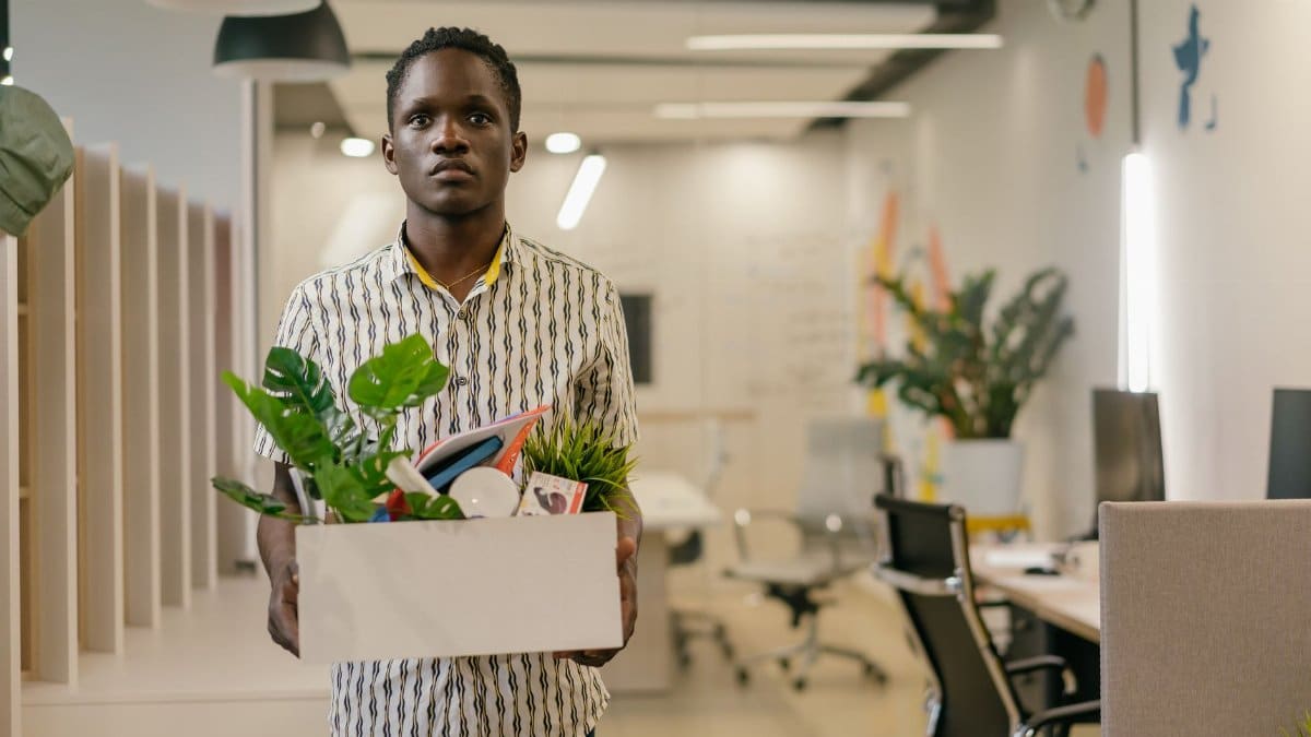 A serious man carries a box of belongings in an office setting, symbolizing dismissal or job change.