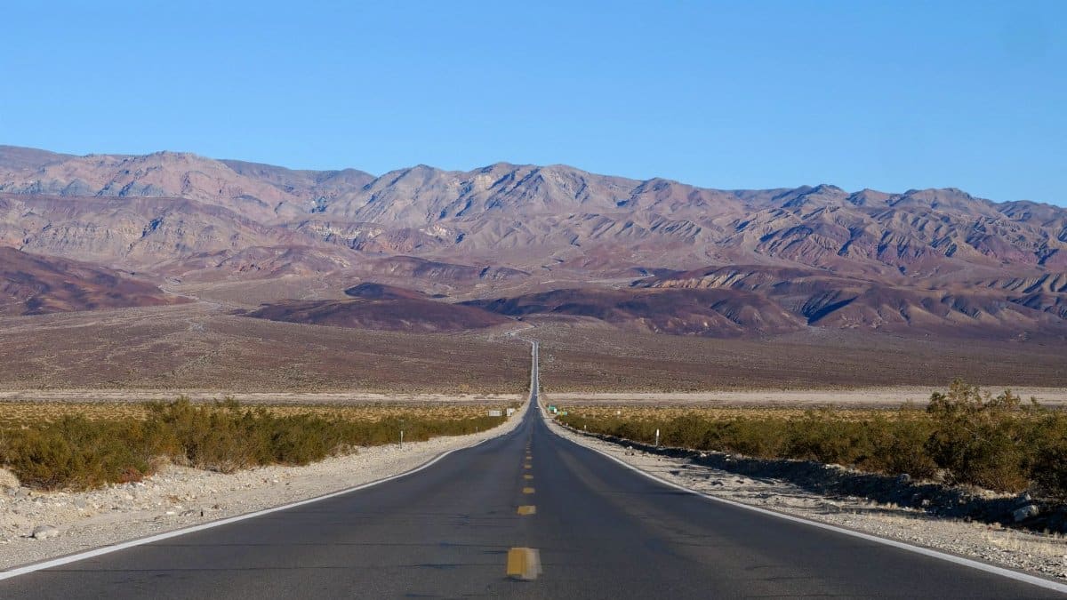 Long stretch of road leading into desert mountains, California, under clear blue skies.