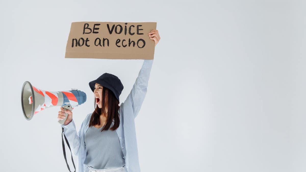 Empowered woman shouts message through megaphone, holding sign that reads 'Be voice, not an echo'.