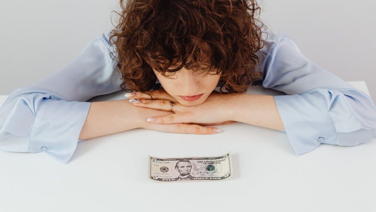 Caucasian woman with curly hair pondering over a five-dollar bill, expressing financial worry.