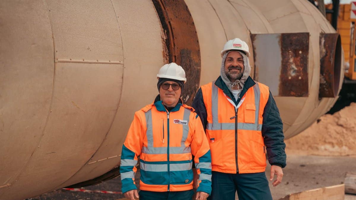 Two construction workers in safety gear standing by industrial machinery on a construction site.