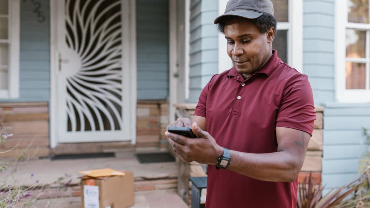 A courier in a maroon shirt checks delivery details on his phone at a residential porch with parcels.