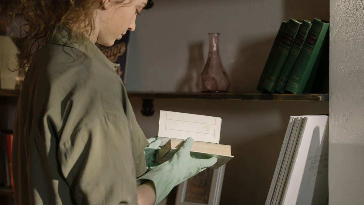 A person wearing gloves organizes books on wooden shelves in a cozy home interior.