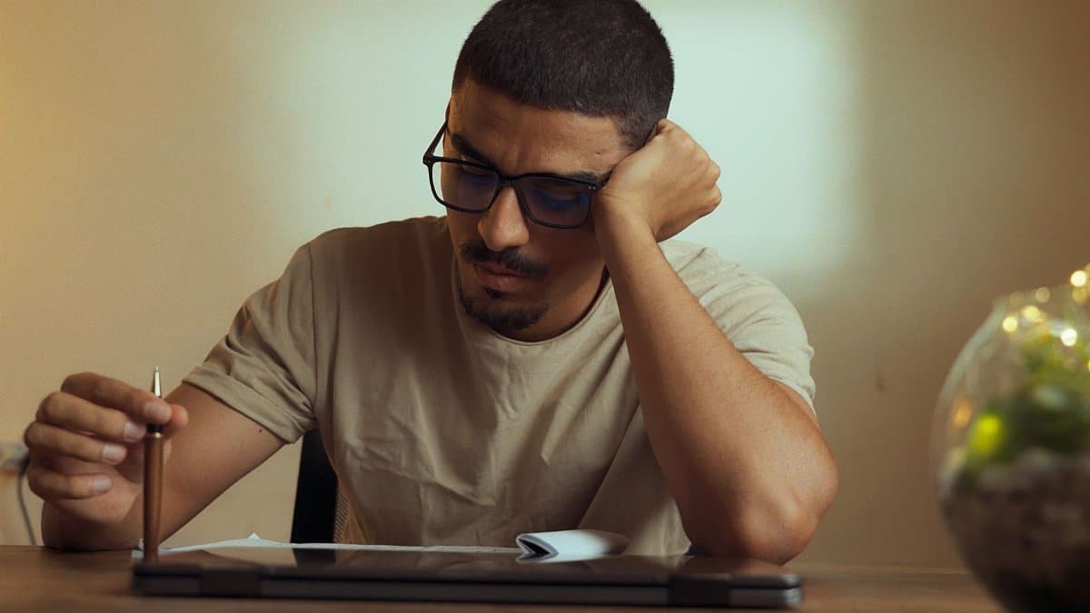 A young man with glasses sits indoors, appearing deep in thought while holding a pencil.