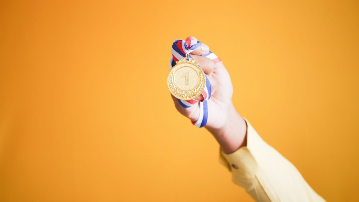 Close-up of hand holding a gold medal with a red, white, and blue ribbon against a vibrant orange background.