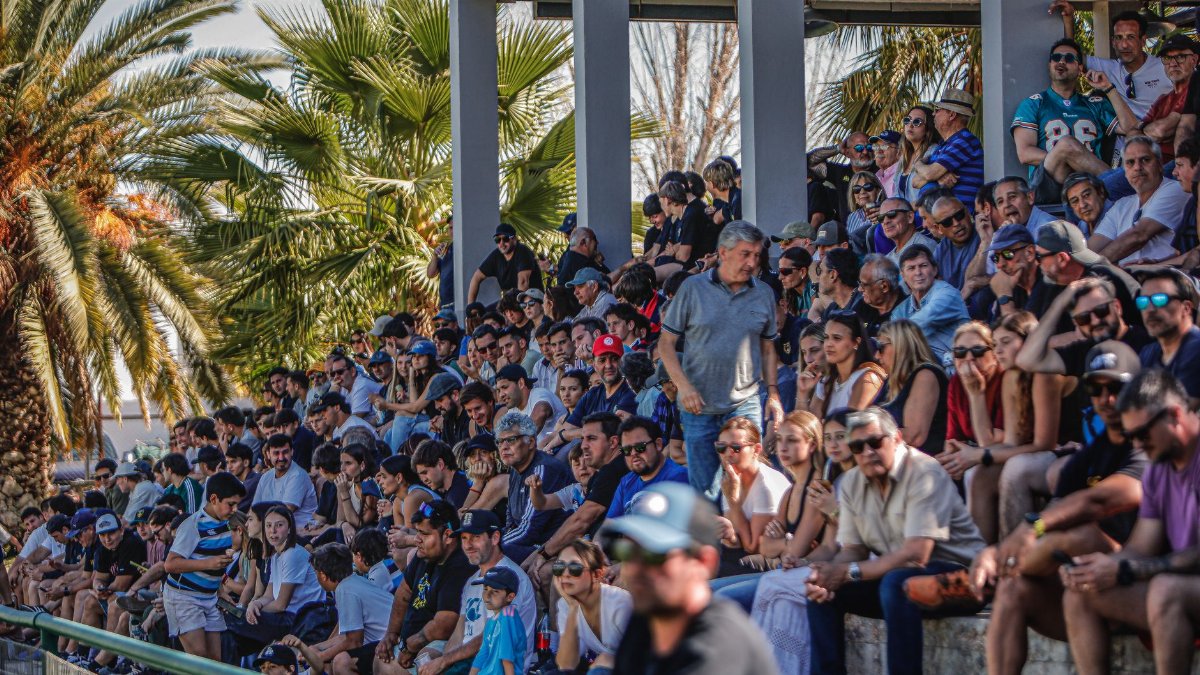 Large diverse crowd seated at an outdoor event, enjoying a sunny day under palm trees.