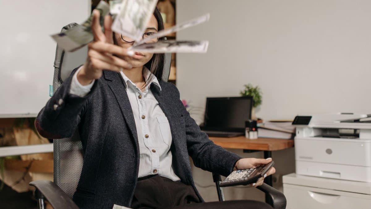 A businesswoman in a suit throws money while calculating finances in a modern office.