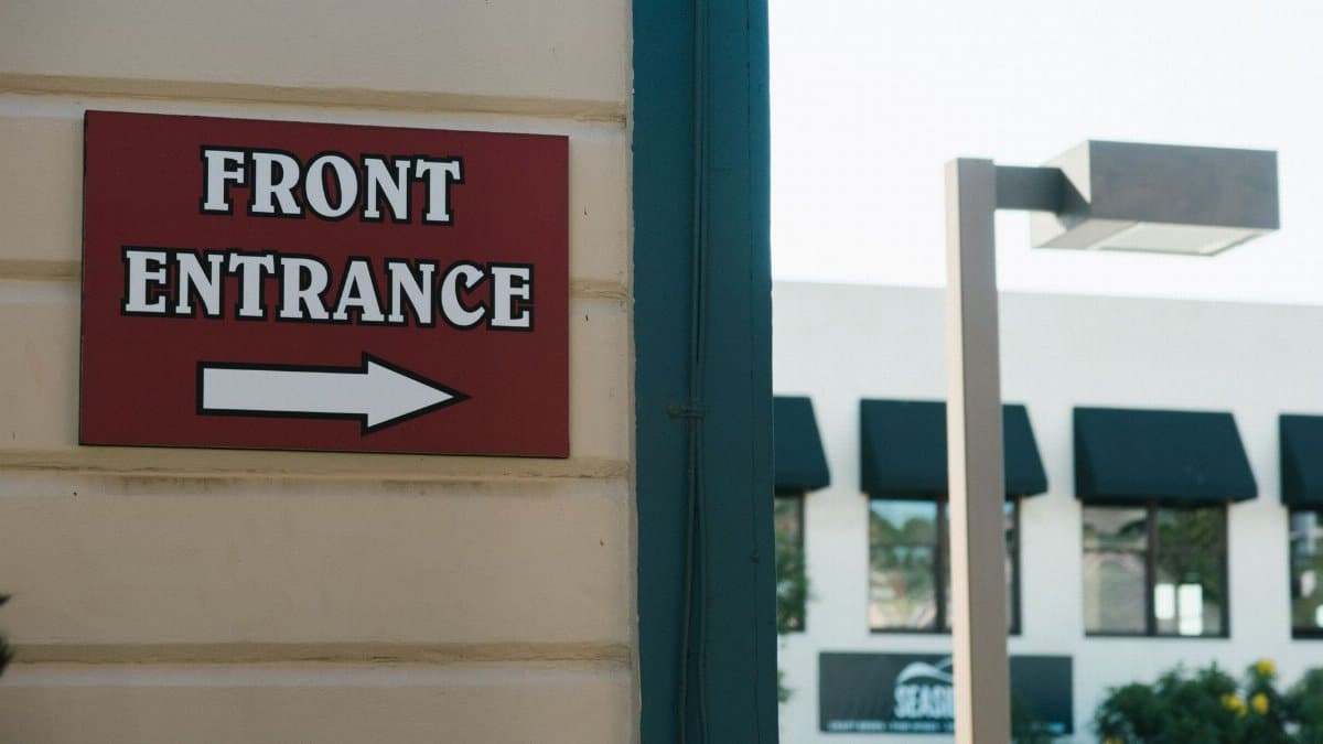 Front entrance sign with arrow indicating direction in urban outdoor setting.