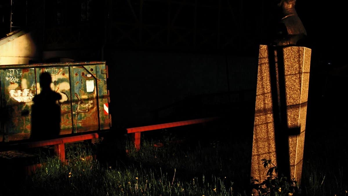 Silhouetted figure casts shadow against a bust in an urban setting at dusk.