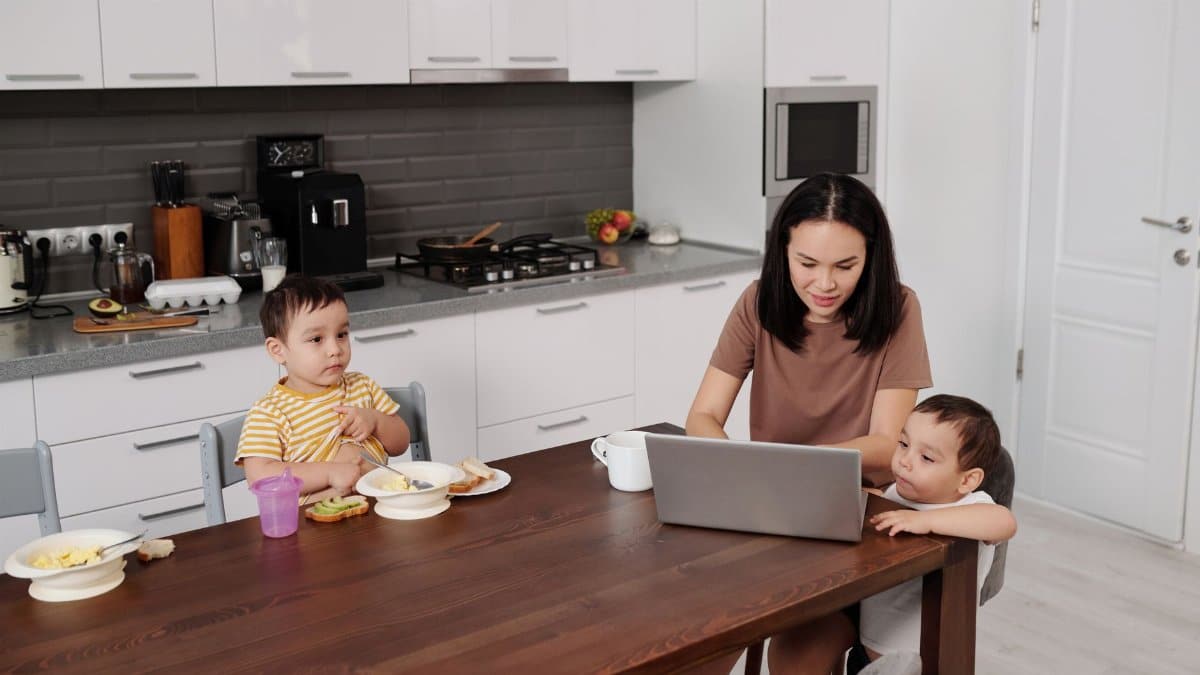 A mother multitasking with her twin sons at the kitchen table while working on a laptop.