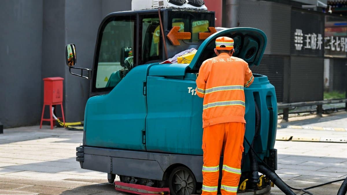 A street cleaner in uniform operates a cleaning machine on an urban sidewalk.