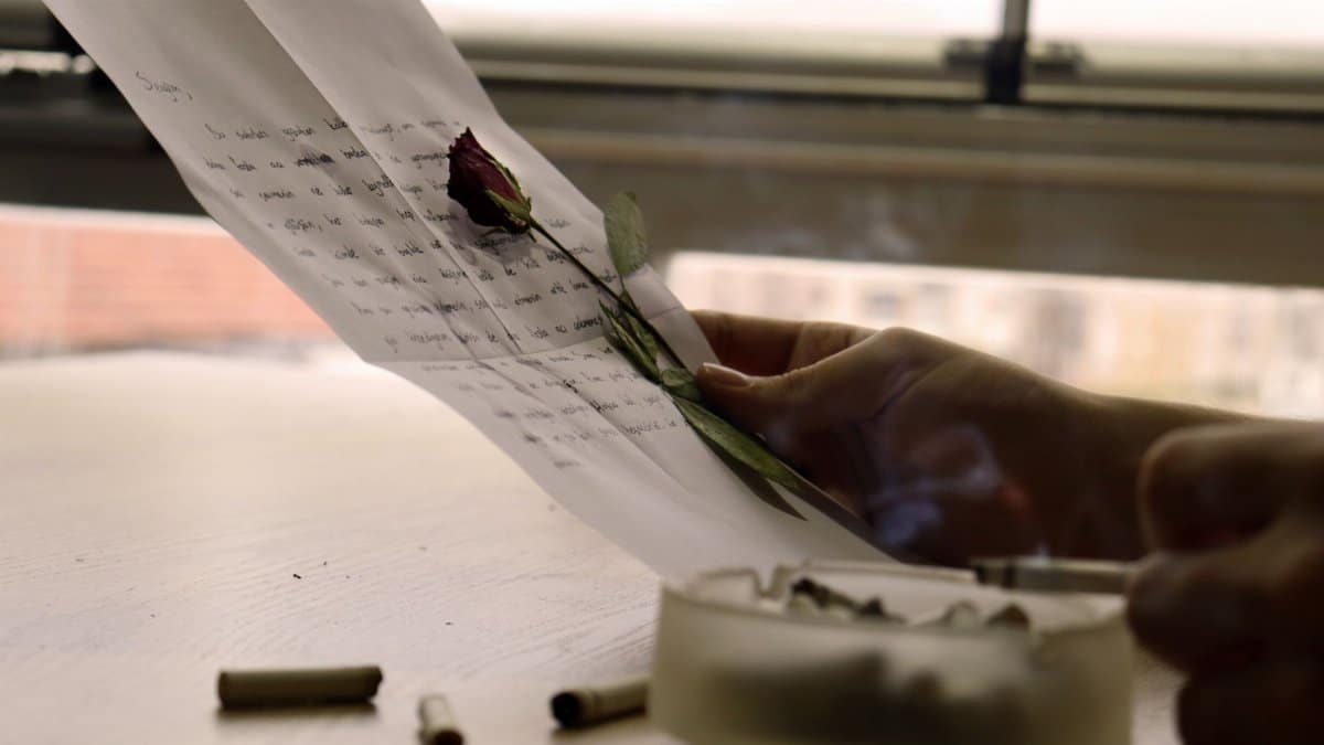 A person holding a letter with a red rose and ashtray; evokes nostalgia and introspection.