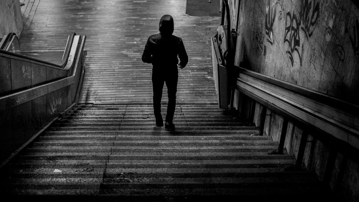 A silhouette of a person wearing a hoodie walking downstairs in a gritty urban environment.