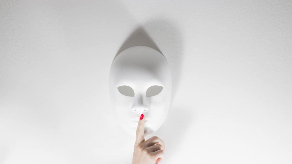A white mask with a human hand gesturing silence on a bright white background, creating a mysterious vibe.