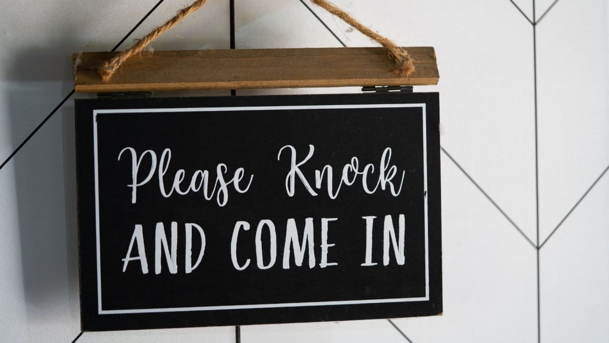 A black, wooden door sign reading please knock and come in.
