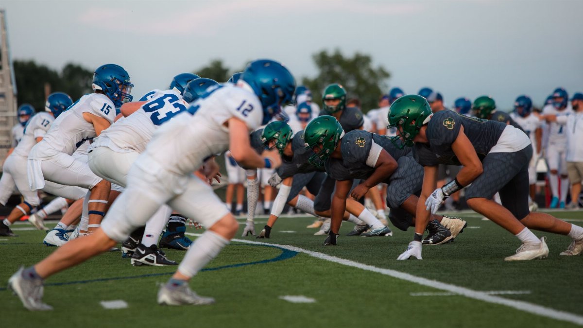 Players in blue and green uniforms face off at the line of scrimmage during a competitive football match.