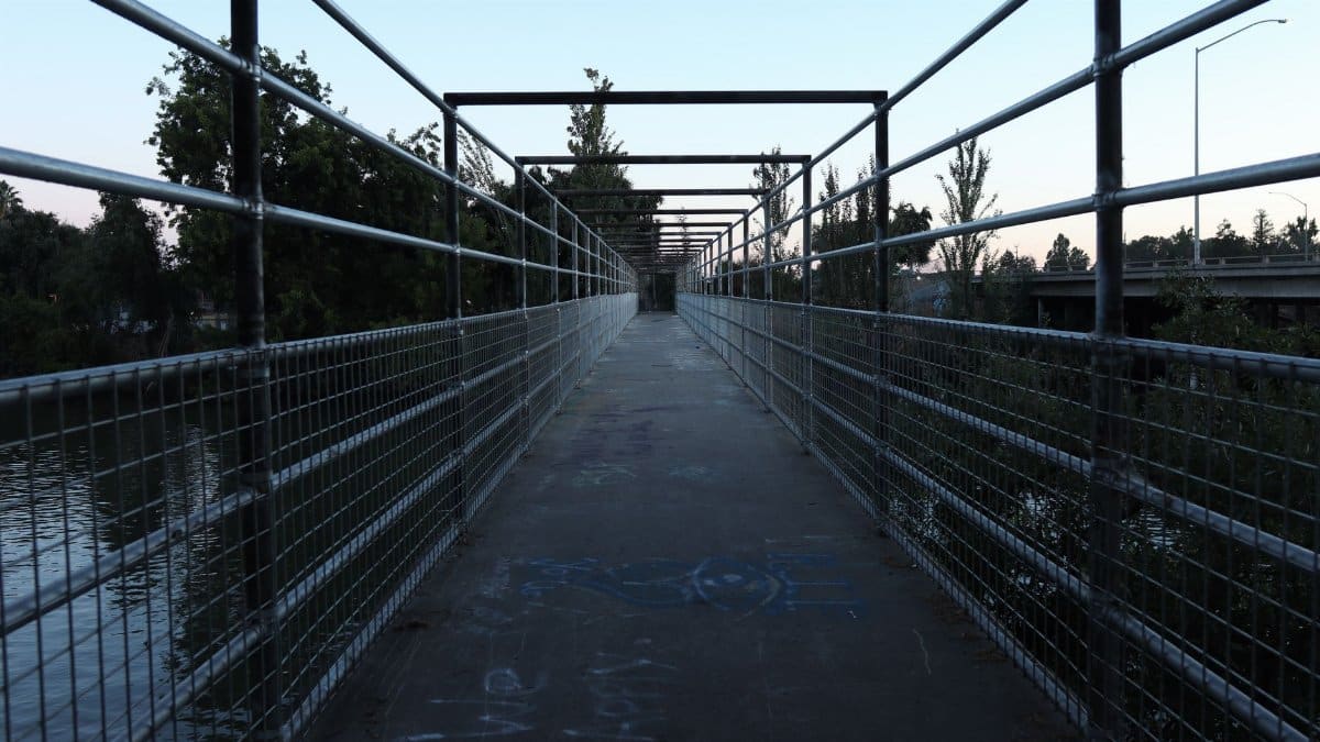 A steel footbridge stretches over a river with trees and a distant highway, captured at dusk.