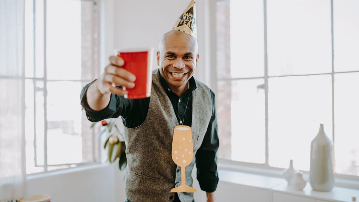 Happy man celebrating New Year indoors with a red cup and party hat.