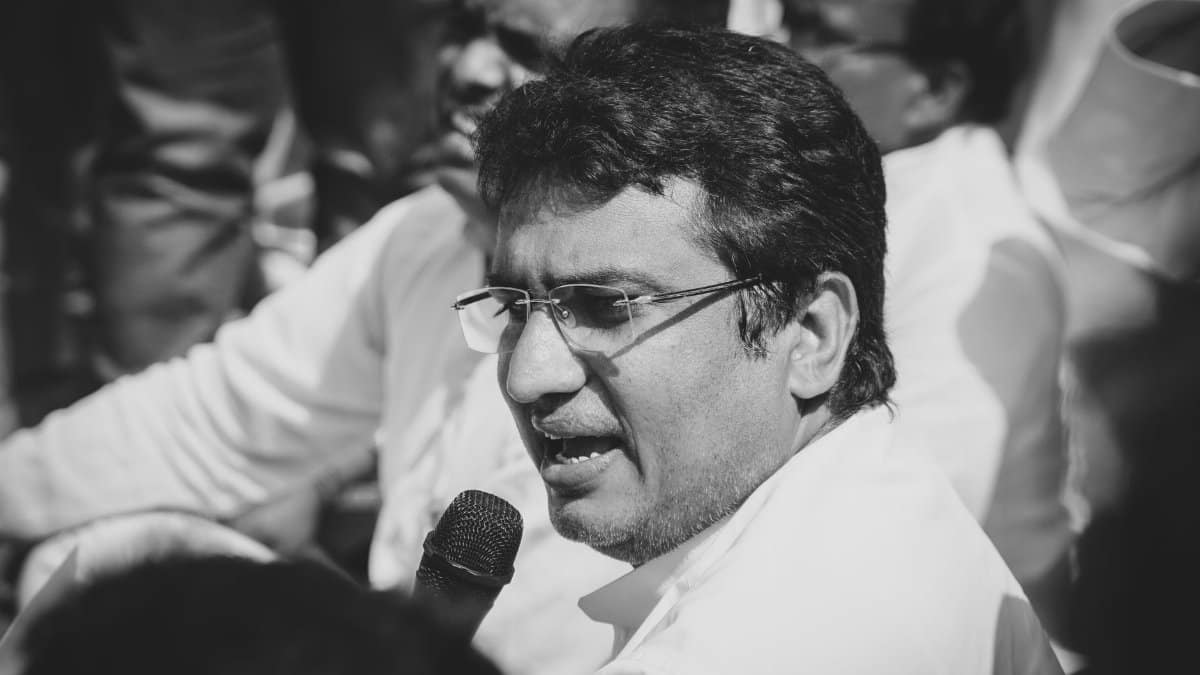Black and white image of a man with eyeglasses speaking into a microphone at an event.
