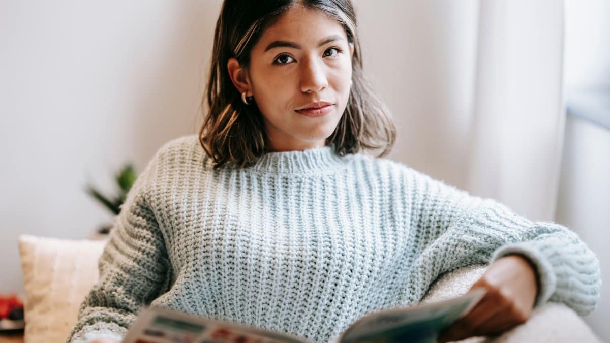 Charming ethnic woman in knitted sweater attentively looking at camera while reading magazine and sitting on couch in white room