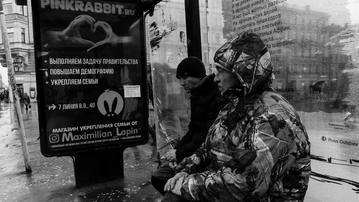 Black and white side view of sad thoughtful exhausted males wearing jacket with hood and winter hat and waiting bus on bench in rainy autumn