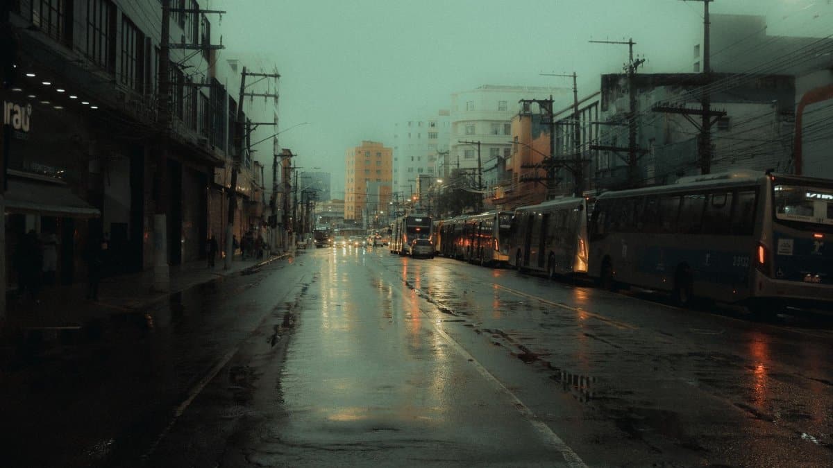 A moody, wet city street with buses lined up under dim lighting, reflecting rain-soaked pavement.